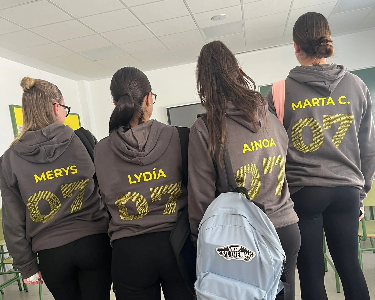 Grupo de espaldas con sudaderas marrones de promoción, nombres y número 07 en amarillo.